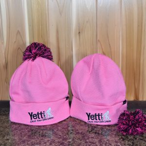 Two pink winter hats with a black Yetti Fish House logo. One hat shows with a pink and black pom on the top of the hat and the other has the pom removed.